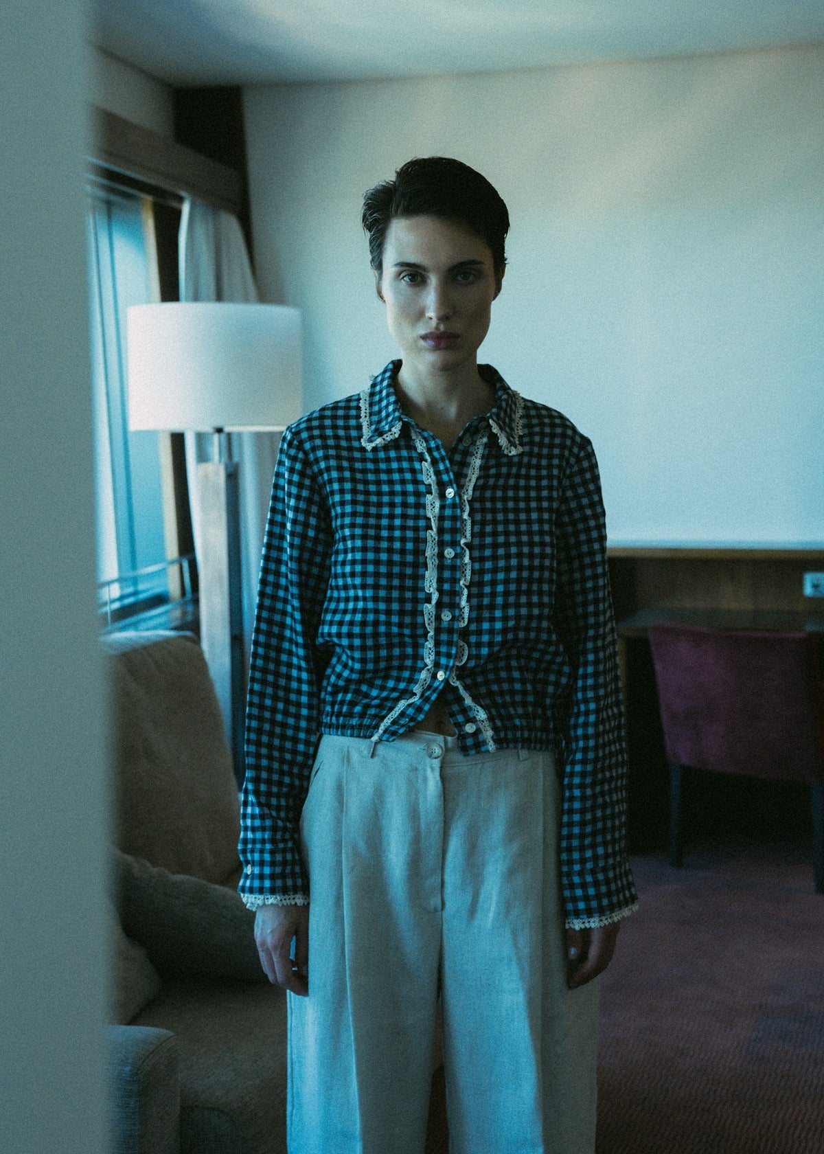 Person wearing a checkered shirt and white pants in a room with a lamp and window.
