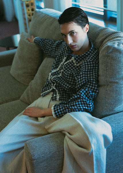 Person wearing a checkered shirt and white pants in a room with a lamp and window.