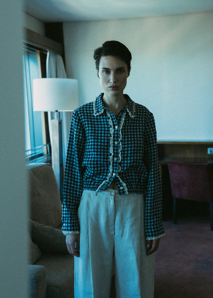 Person wearing a checkered shirt and white pants in a room with a lamp and window.