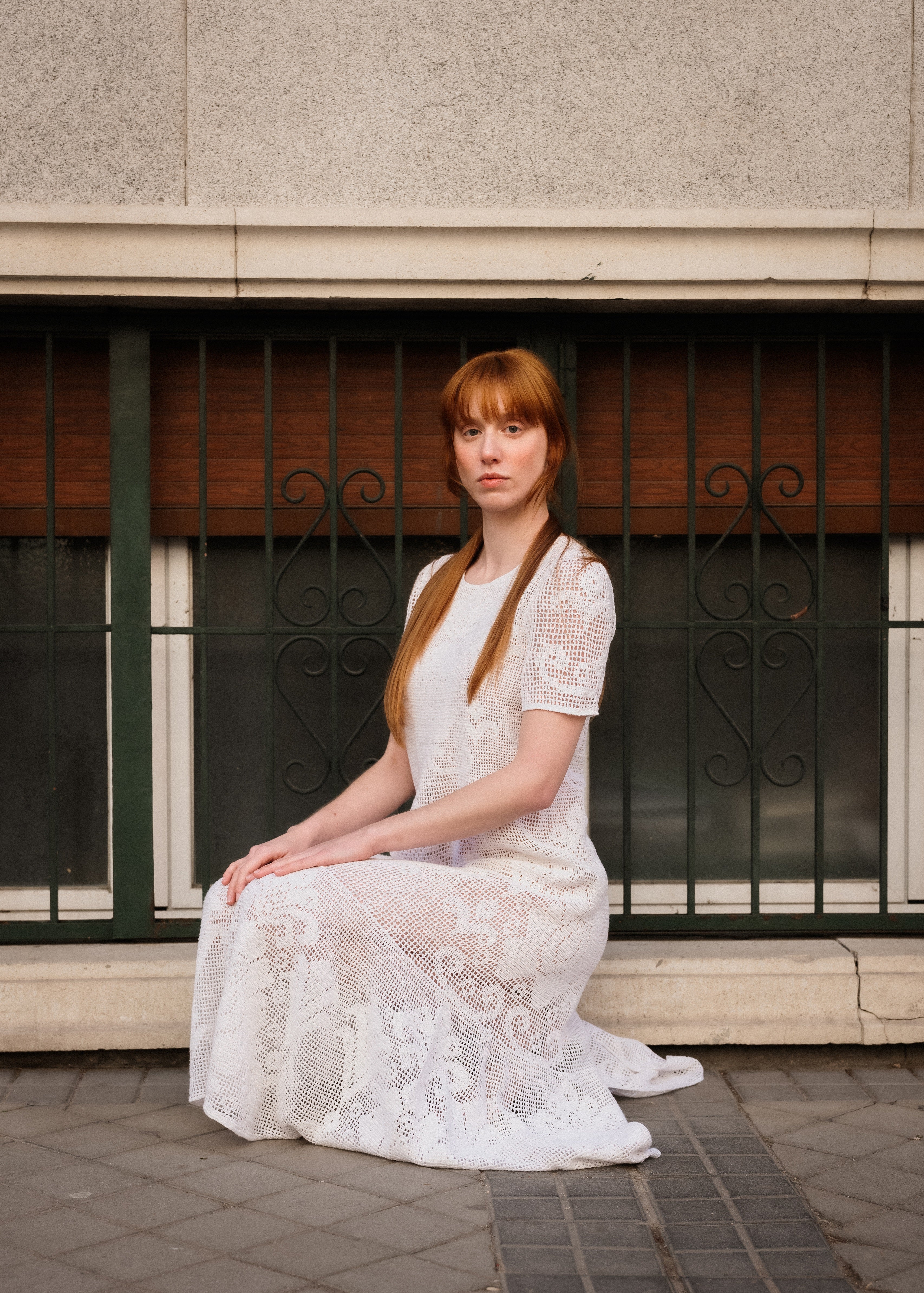Woman in a white lace dress sitting on a sidewalk with a building in the background