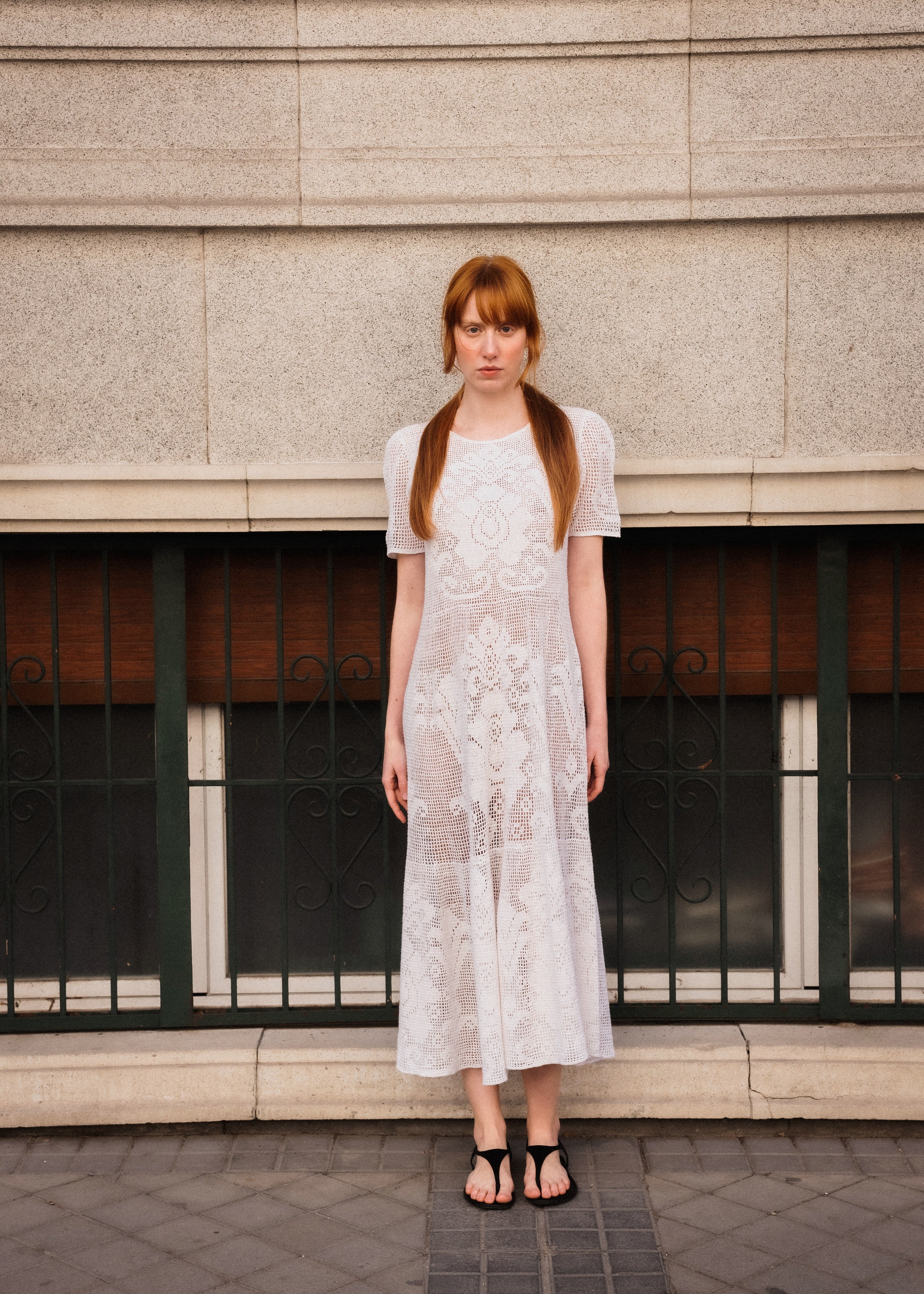 Woman in a white lace dress standing in front of a building with a window.