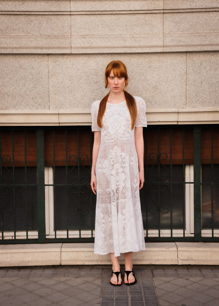Woman in a white lace dress standing in front of a building with a window.
