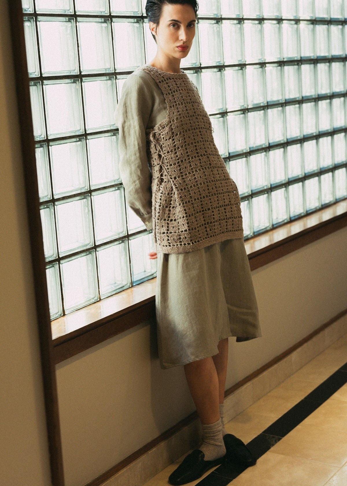 Woman wearing a beige crochet top and skirt standing in front of a glass paneled wall.