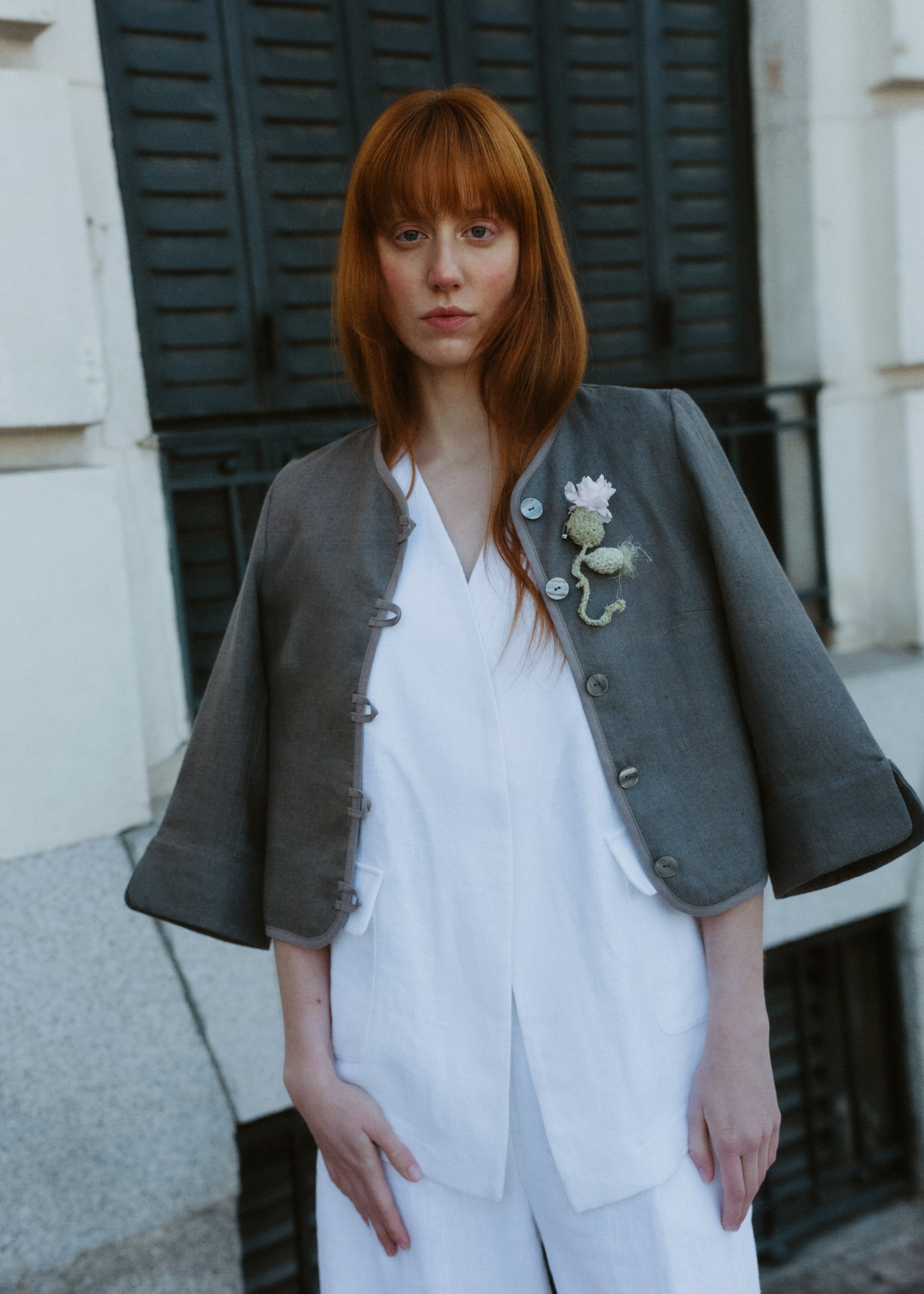 Woman wearing a gray blazer over a white outfit with a floral brooch, standing outdoors.