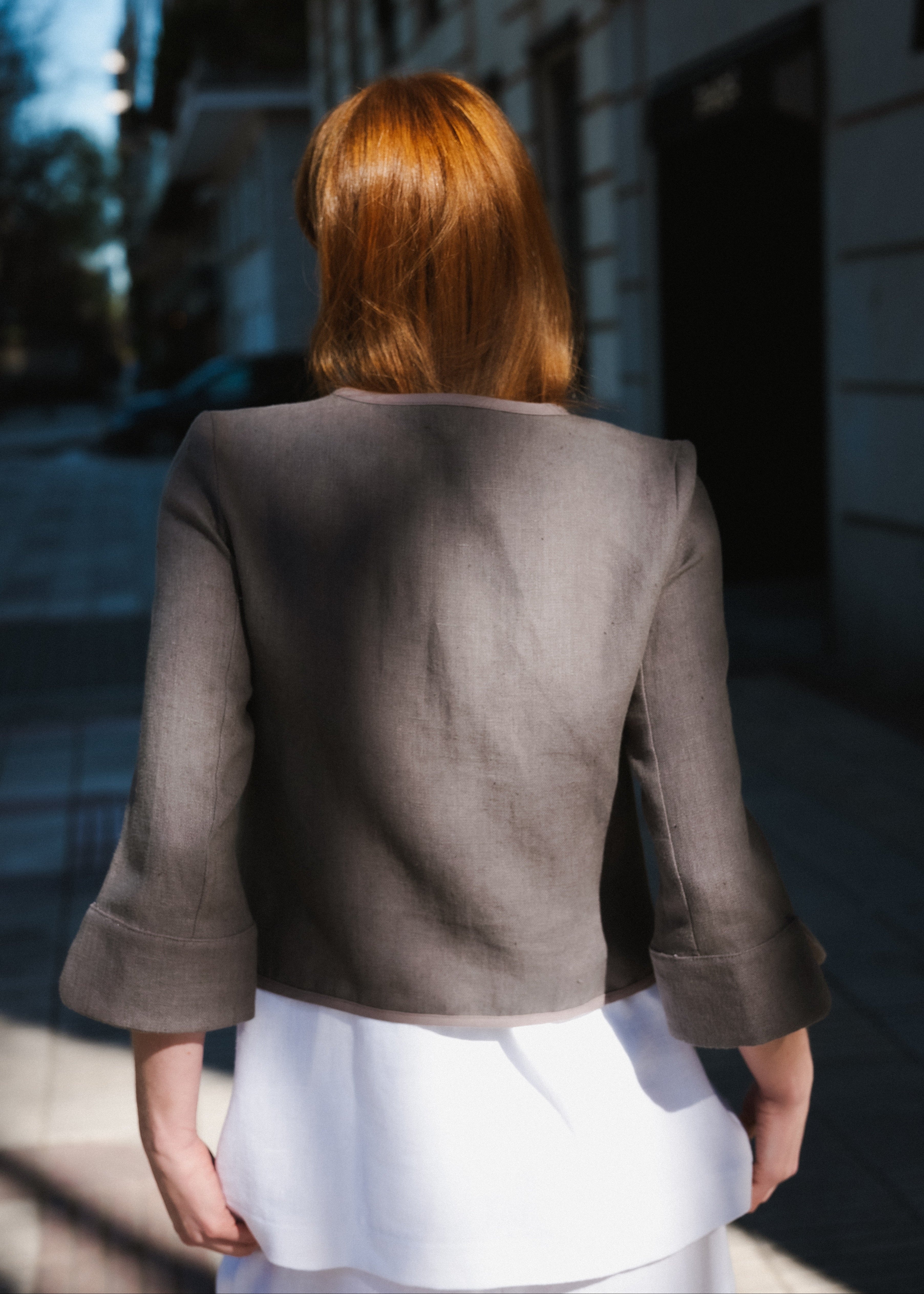 Person wearing a brown blazer and white skirt walking away from the camera on a city street.