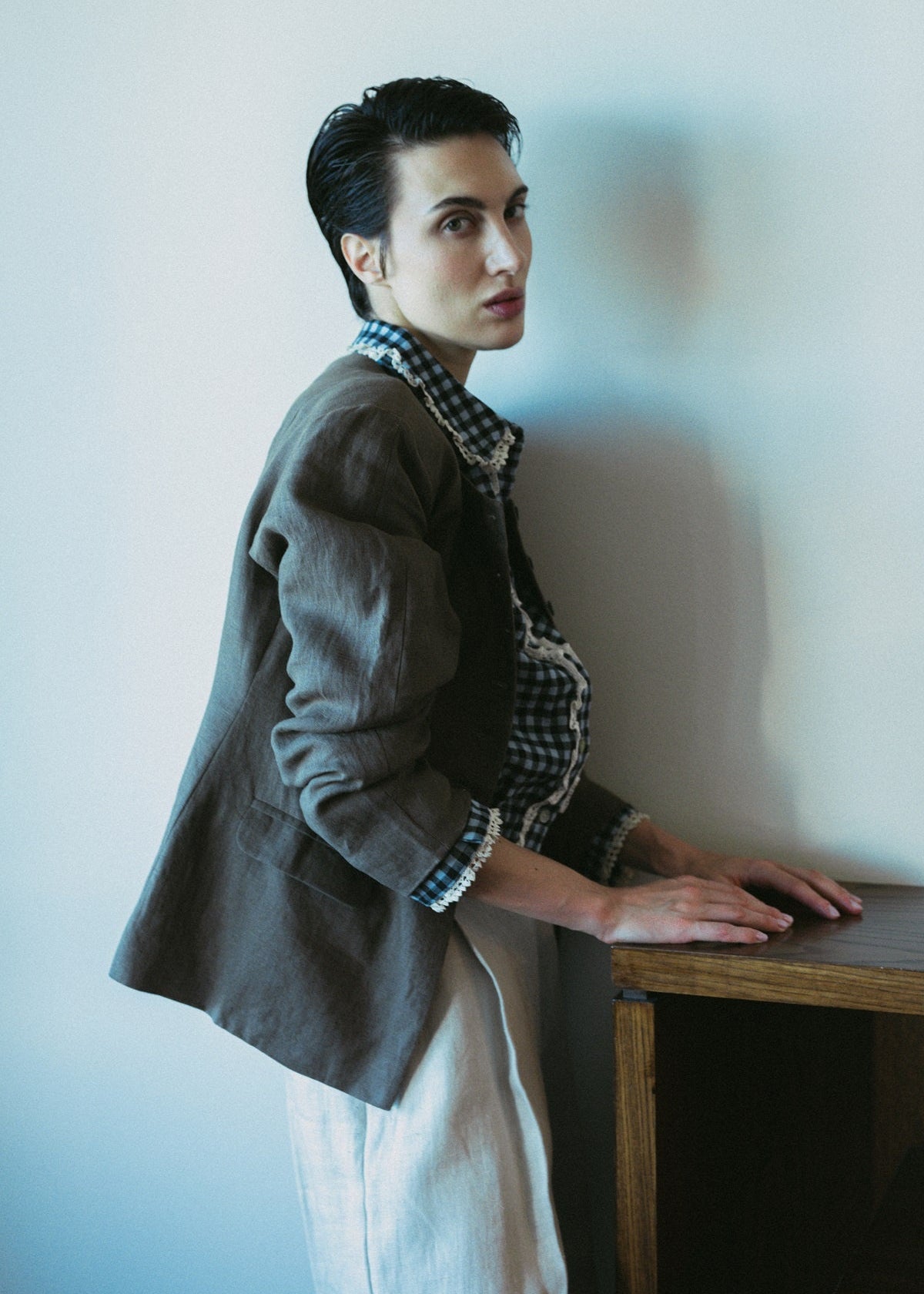 Person wearing a gray blazer and plaid shirt leaning against a white wall.