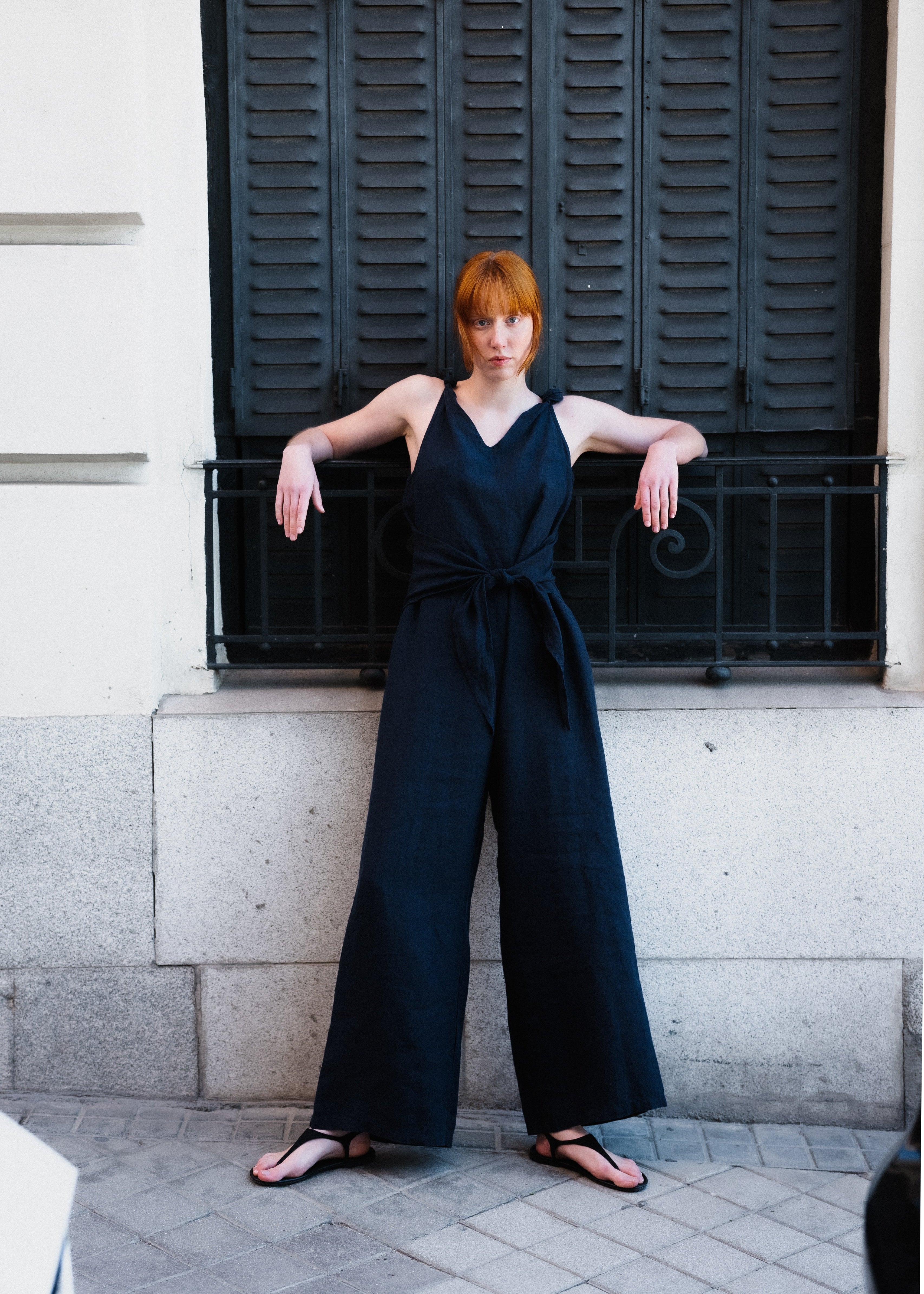Woman wearing a dark blue jumpsuit standing against a building with black shutters.