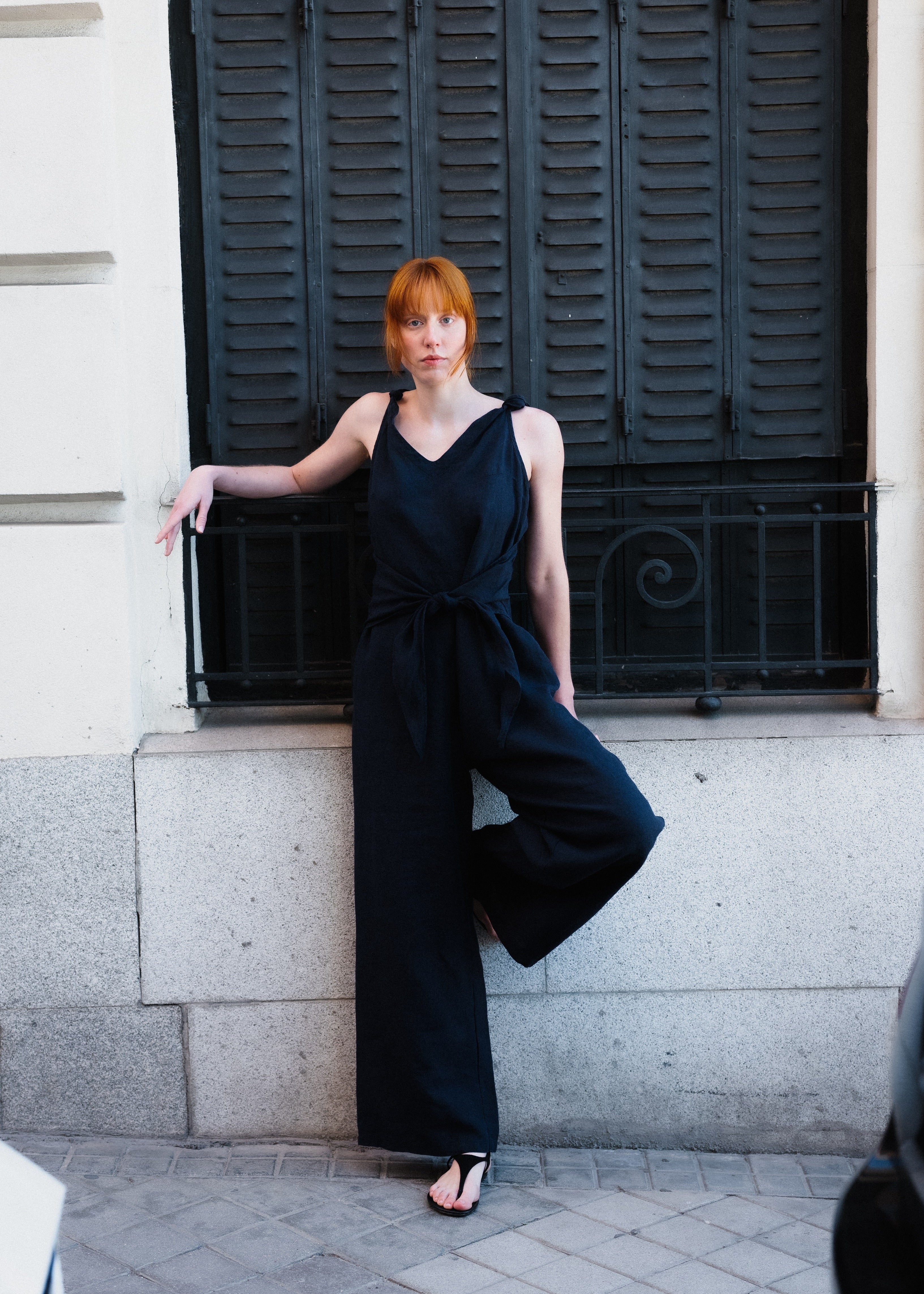 Woman in a black jumpsuit standing against a building with black shutters.