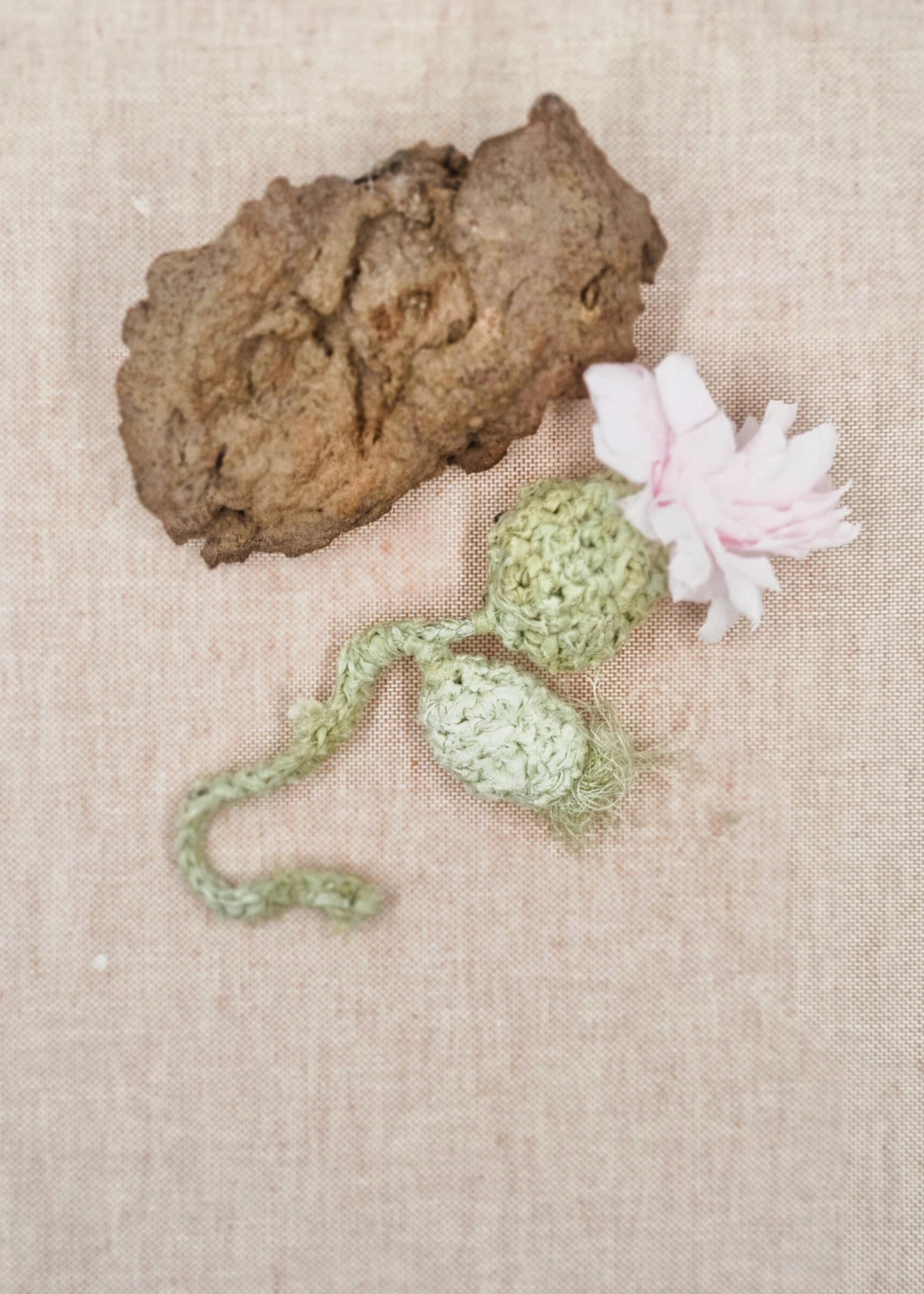 Small rock, green crocheted plant, and pink flower on a beige fabric background