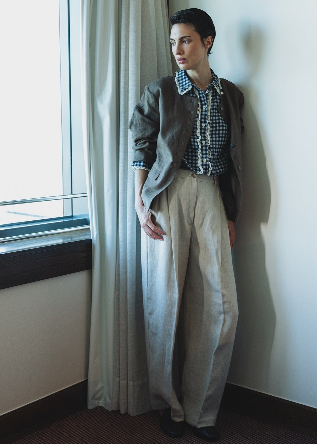 Person standing by a window with light curtains, wearing a patterned shirt and wide-leg pants.