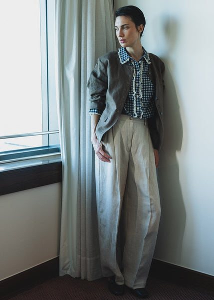 Person standing by a window with light curtains, wearing a patterned shirt and wide-leg pants.