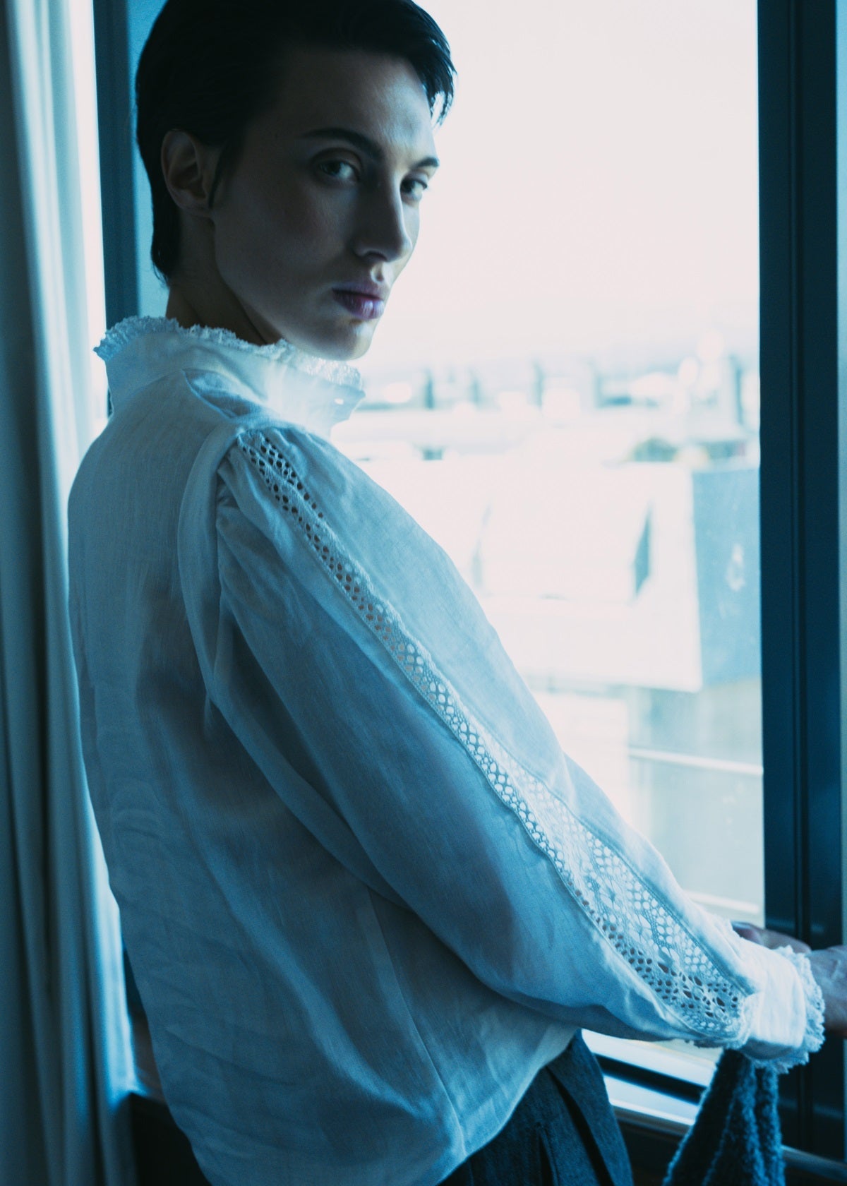 Person wearing a white lace-trimmed shirt by a window with a blurred background