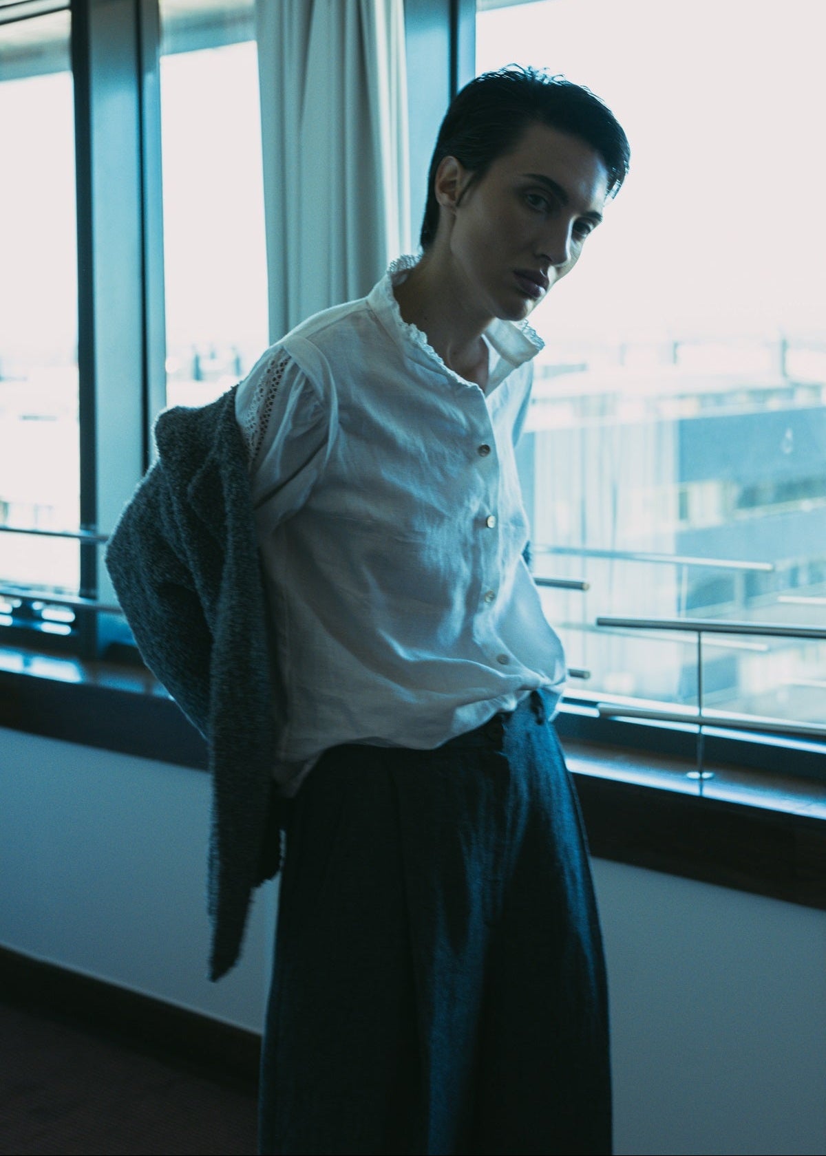 Person wearing a white shirt and dark pants standing by a window with a cityscape view.